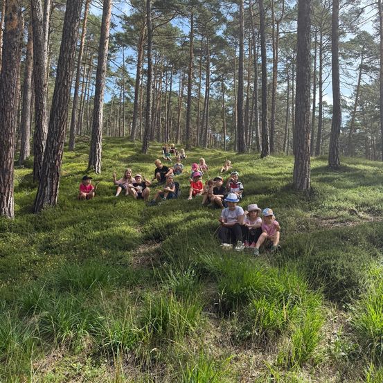 Eine Gruppe von Kindern und Erwachsenen sitzt in einem Wald, einige tragen Hüte und Schuhe, mit hohen Bäumen um sie herum.
