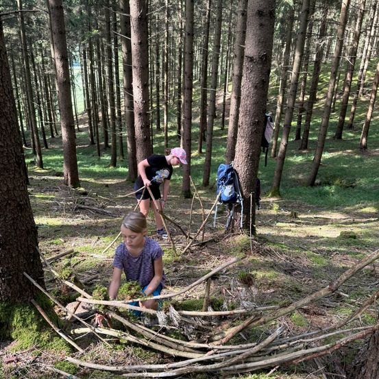 Zwei junge Mädchen sind in einem Wald, eine beugt sich vor, um Stöcke aufzuheben, und die andere hält einen Stock und einen Hut.
