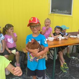 Ein Junge in einem Ferrari-Hut hält ein Huhn, während Kinder hinter ihm an einem Tisch sitzen.
