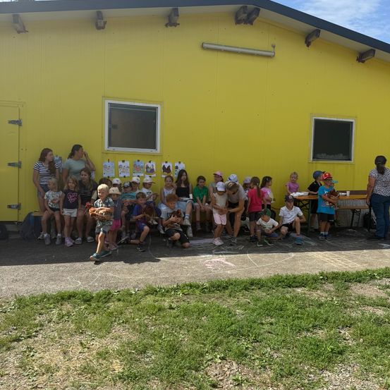 Eine Gruppe von Kindern und Erwachsenen versammelt sich vor einem gelben Gebäude, einige halten Baseballhandschuhe.