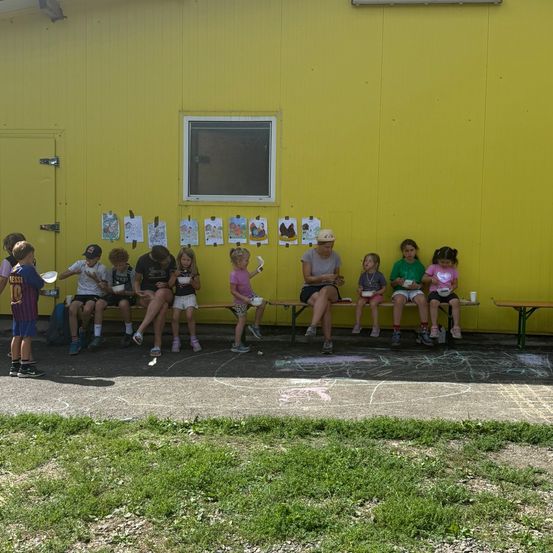 Eine Gruppe von Kindern und Erwachsenen sitzt auf Bänken vor einem gelben Gebäude, einige halten Teller und Tassen. Zeichnungen sind an der Wand befestigt.