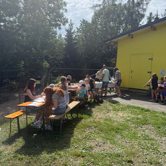 Eine Gruppe von Menschen sitzt an Picknicktischen im Freien, einige stehen und andere gehen herum. Im Hintergrund befindet sich ein gelbes Gebäude.