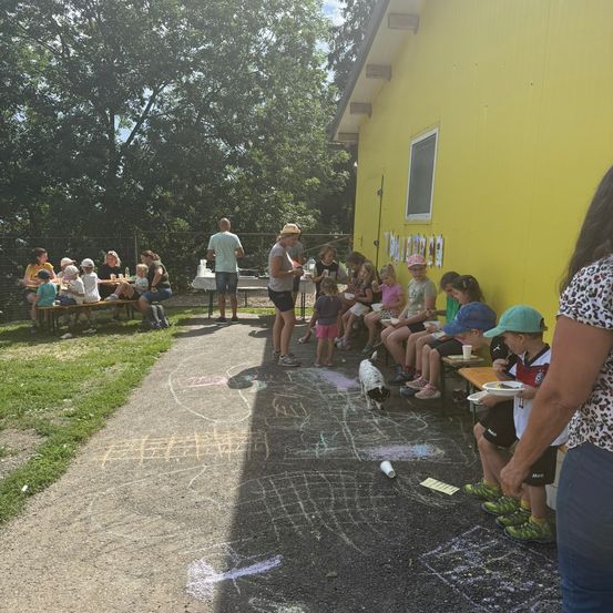 Eine Gruppe von Kindern und Erwachsenen sitzt auf Bänken vor einem gelben Gebäude, einige essen, andere stehen. Ein Hund läuft in der Nähe.