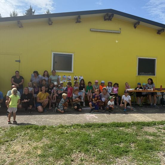 Eine Gruppe von Menschen, einschließlich Kindern, versammelt sich vor einem gelben Gebäude, einige sitzen auf dem Boden und andere stehen. Es gibt Tische mit Essen und ein Grasfeld in der Nähe.