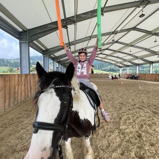Ein junges Mädchen mit Helm reitet ein schwarz-weißes Pferd in einer überdachten Reithalle. Sie hält einen grünen Stab mit beiden Händen.