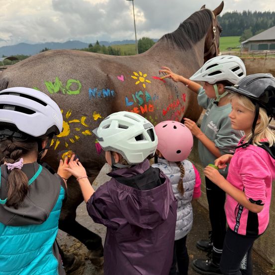 Eine Gruppe junger Kinder mit Helmen malt ein Pferd mit verschiedenen farbenfrohen Zeichnungen und Wörtern.