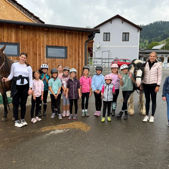 Eine Gruppe von Kindern und zwei Frauen steht vor einem Holzgebäude mit einem Pferd und einem Pony. Die Kinder tragen Helme und Jacken.