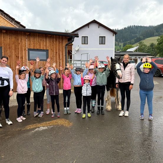 Eine Gruppe von Kindern und Erwachsenen in Helmen und Turnschuhen posiert für ein Foto vor einer Scheune mit einem Pferd.