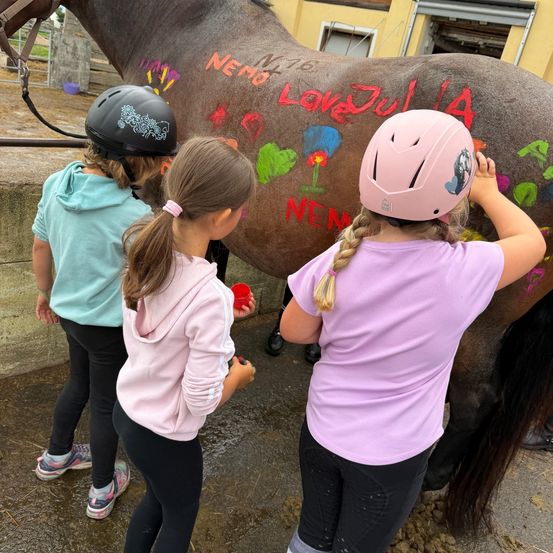 Drei junge Mädchen in rosa Helmen schreiben auf einem Pferd mit bunten Zeichnungen und Wörtern wie Nemo und Love Julia. Das Pferd steht vor einem Gebäude.