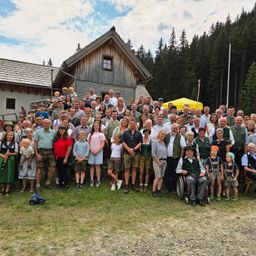 Eine große Gruppe von Menschen, einschließlich Kindern und Erwachsenen, versammelt sich vor einem Holzhaus in einem Berggebiet. Einige tragen traditionelle Kleidung.