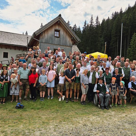Eine große Gruppe von Menschen, einschließlich Kindern und Erwachsenen, versammelt sich vor einem Holzhaus in einem Berggebiet. Einige tragen traditionelle Kleidung.