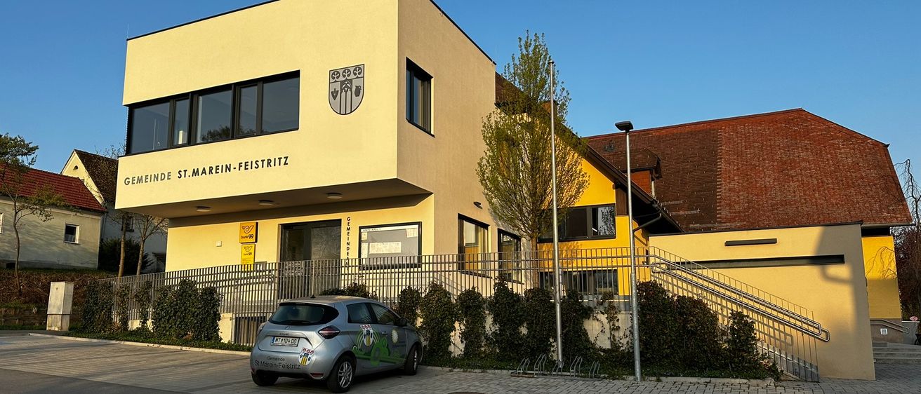 Ein silberner Wagen ist vor einem gelben Gebäude mit einem Logo über dem Eingang geparkt. Das Gebäude hat Glasfenster und eine Treppe an der Seite.