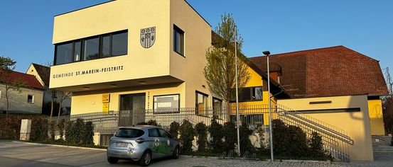 Ein silberner Wagen ist vor einem gelben Gebäude mit einem Logo über dem Eingang geparkt. Das Gebäude hat Glasfenster und eine Treppe an der Seite.