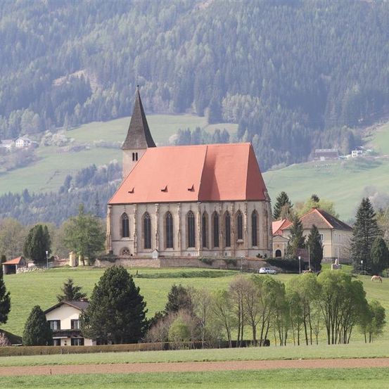 Eine Kirche mit rotem Dach steht auf einem Hügel, umgeben von grünen Feldern und Bäumen. Sie hat einen Turm und mehrere Fenster. In der Nähe sind ein Haus und ein geparktes Auto zu sehen.