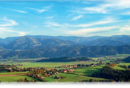 Ein friedliches Tal mit sanften Hügeln und Bergen in der Ferne, üppige grüne Felder und ein kleines Dorf, das zwischen Bäumen eingebettet ist. Der Himmel ist blau mit flaumigen Wolken.