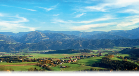 Ein friedliches Tal mit sanften Hügeln und Bergen in der Ferne, üppige grüne Felder und ein kleines Dorf, das zwischen Bäumen eingebettet ist. Der Himmel ist blau mit flaumigen Wolken.