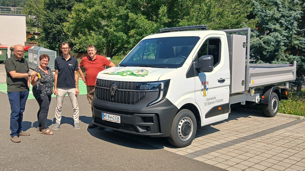 Two men stand beside a white Renault truck with green and yellow graphics on the front, parked on a paved area with trees in the background.