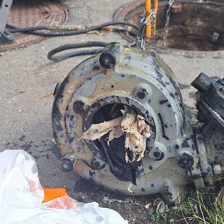 A damaged mechanical device with exposed internal parts and debris, possibly from a sewer system, lying on the ground near a manhole cover.