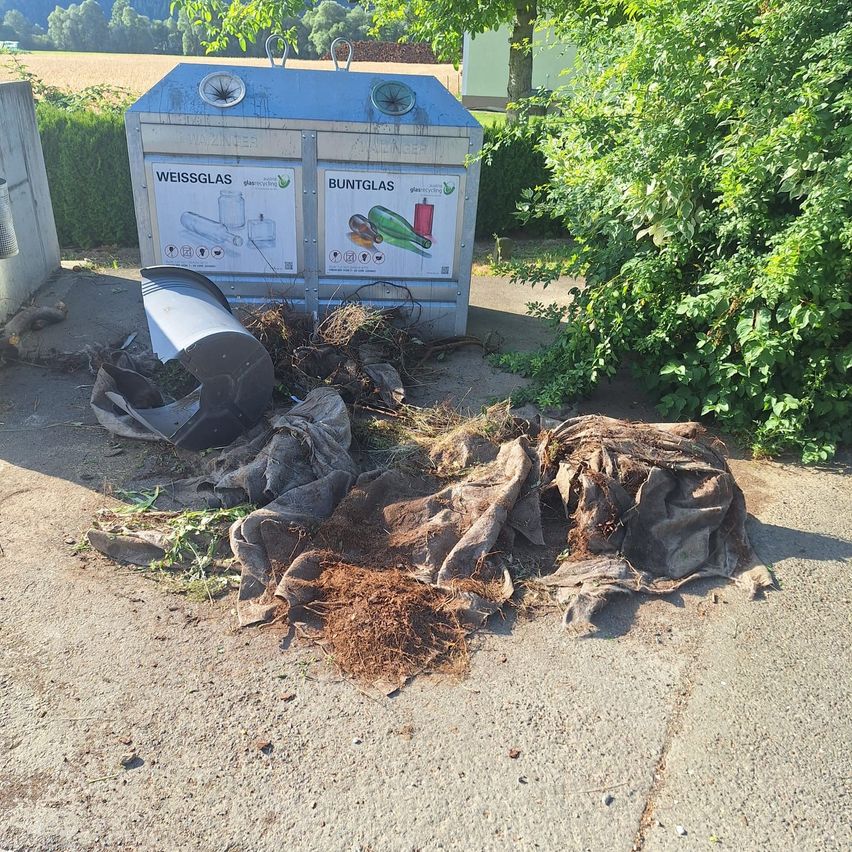 Ein Recyclingbehälter mit Schildern für weißes und farbiges Glas liegt umgekippt auf dem Boden, mit umherliegenden Abfällen.