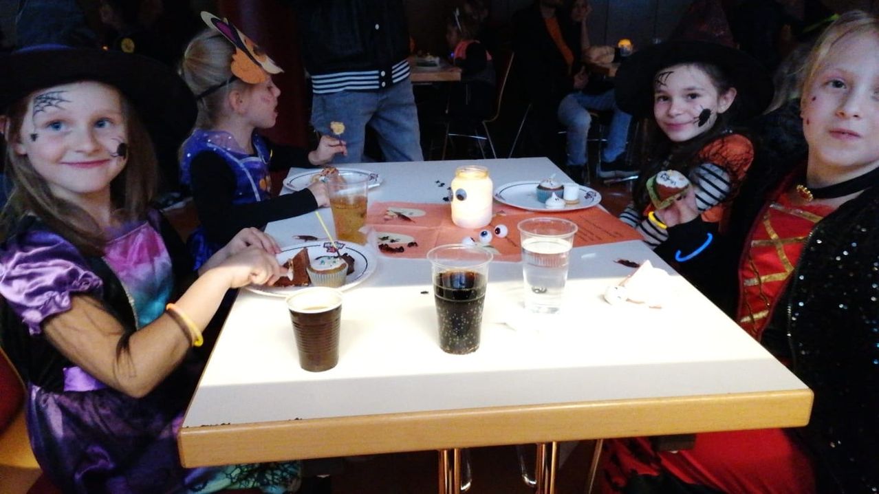 Several children are sitting at a table with cupcakes, drinks, and a candle. One child has a Halloween costume and a cupcake on a plate.
