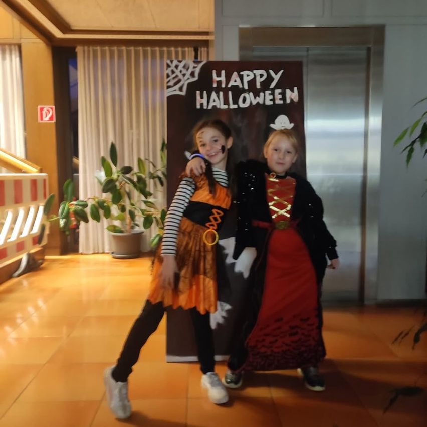 Two children dressed in Halloween costumes stand before a 'Happy Halloween' banner, surrounded by indoor plants and an elevator.