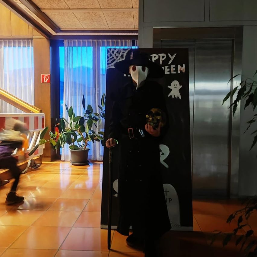 A person dressed as a plague doctor stands in a hall, holding a skull and cane, with Halloween decorations in the background.
