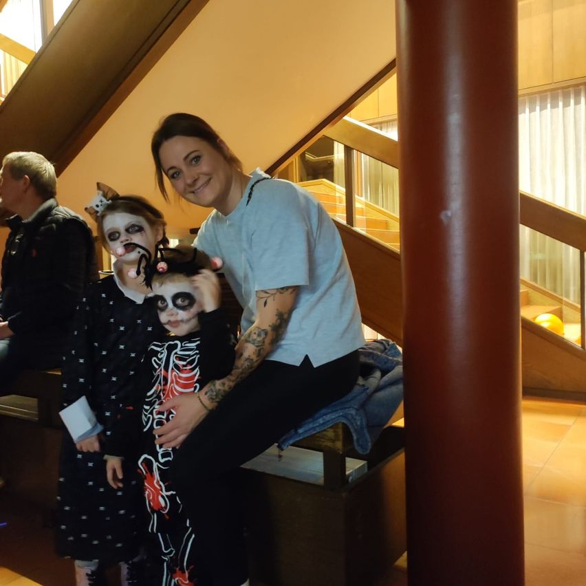 A woman and two children in costumes are smiling for a photo. They are sitting on a bench. Behind them, a staircase leads up to a hallway.