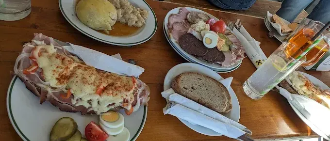 Ein hölzerner Tisch zeigt verschiedene Speisen: ein Teller mit einem Fleischgericht, ein Fleischsandwich, ein Teller mit Gemüse und Eiern und ein Glas Wasser.
