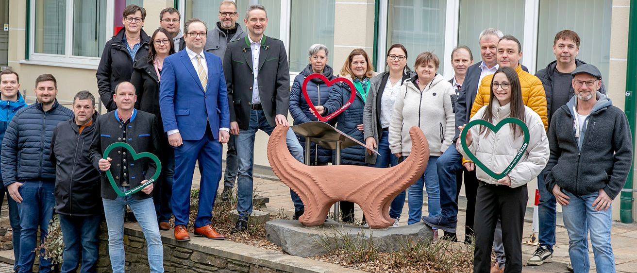 Eine Gruppe von Menschen, darunter ein Mann in einem blauen Anzug, steht vor einem gelben Gebäude mit einer Skulptur und herzförmigen Rahmen.