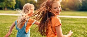 Zwei junge Mädchen rennen fröhlich auf einer grünen Wiese, ihr Haar weht im Wind. Die Sonne scheint hell hinter ihnen und schafft eine warme und lebendige Atmosphäre.