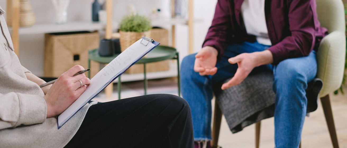 Eine Person sitzt und notiert mit einem Stift, während eine andere Person spricht. Ein Schreibtisch und Pflanzen sind im Hintergrund.
