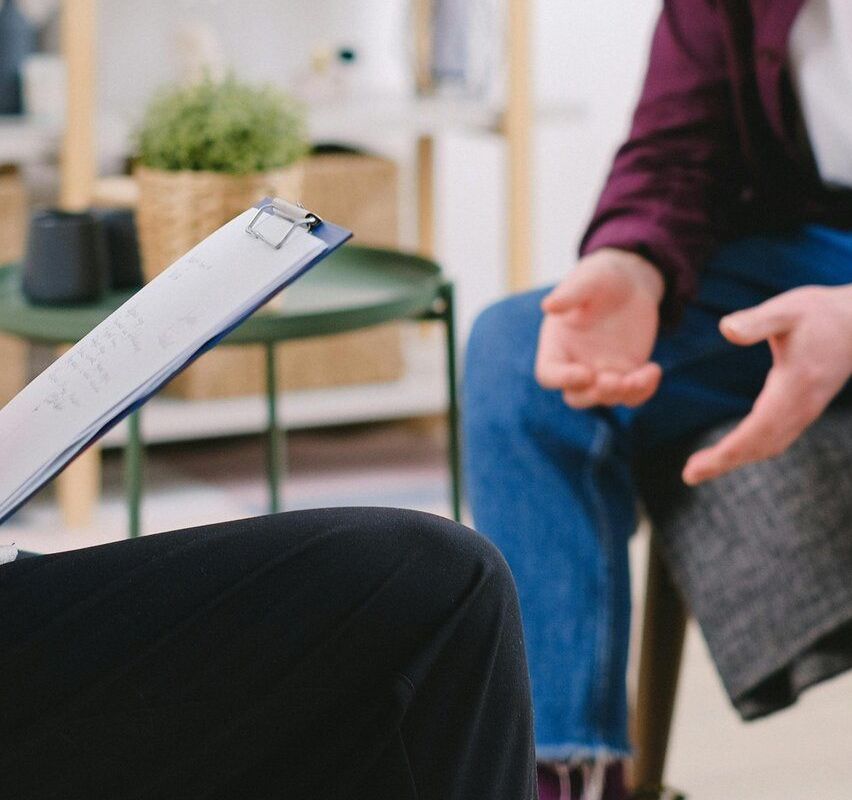 Eine Person sitzt und notiert mit einem Stift, während eine andere Person spricht. Ein Schreibtisch und Pflanzen sind im Hintergrund.