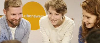 Eine Gruppe von Menschen versammelt sich im Kreis, lächelt und spricht. Eine Frau in Weiß sitzt in der Mitte. Ein gelbes Schild mit Text ist im Hintergrund.