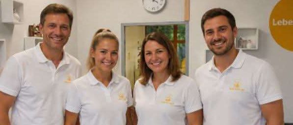 Vier Personen in passenden weißen Shirts und gelben Shorts stehen gemeinsam in einem Büro und lächeln für ein Foto. Eine Uhr und ein Schild mit der Aufschrift 'Lebensweg' sind an der Wand hinter ihnen zu sehen.