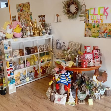 Ein Raum gefüllt mit verschiedenen Dekorationsartikeln, darunter Figuren, Körbe und Blumen, die auf Regalen und einem Tisch angeordnet sind. Ein Kranz und ein Schild mit der Aufschrift 'CLICK COLLET SHOP' hängen an der Wand.