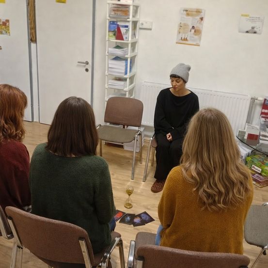 Fünf Frauen sitzen in einem Kreis auf einem Holzboden, eine mit einer Mütze spricht. Sie halten Karten, ein Becher steht auf dem Boden. Dahinter befindet sich ein weißer Regal, ein Heizkörper und ein Glastisch.