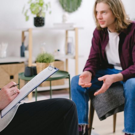 Zwei Menschen sitzen, einer hält ein Klemmbrett und einen Stift, möglicherweise in einer Beratung. In der Nähe stehen eine Pflanze und kleine Gegenstände auf einem Regal.