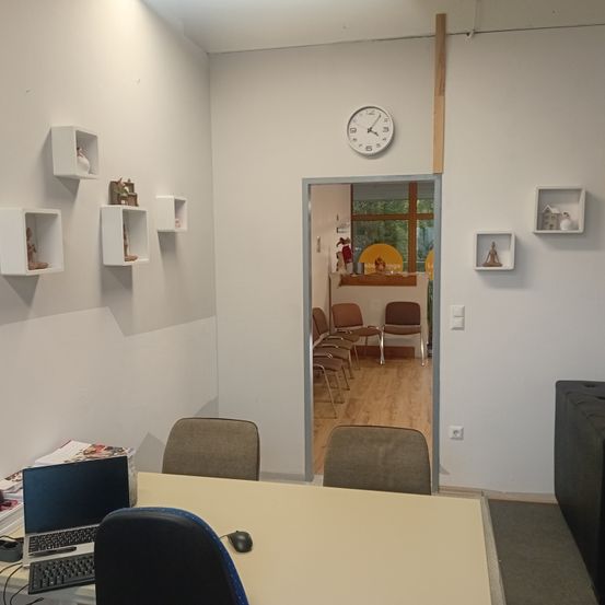 Ein leeres Büro mit einem Schreibtisch, einem Stuhl und einem Laptop. An der Wand befinden sich Regale mit Gegenständen und eine Uhr. Der Raum hat Holzboden und eine offene Tür, die zu einem anderen Raum mit Stühlen führt.