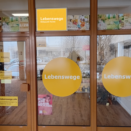 Der Eingang eines Geschafts namens Lebenswege mit einem gelben Schild und großen gelben Kreisen auf den Glastüren. Es gibt zwei Schilder mit Kontaktdaten auf der linken Tür.