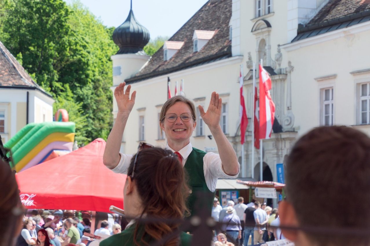Eine Frau mit Brille und grüner Weste hebt ihre Hände vor einem Gebäude mit Fahnen und einer Menschenmenge.