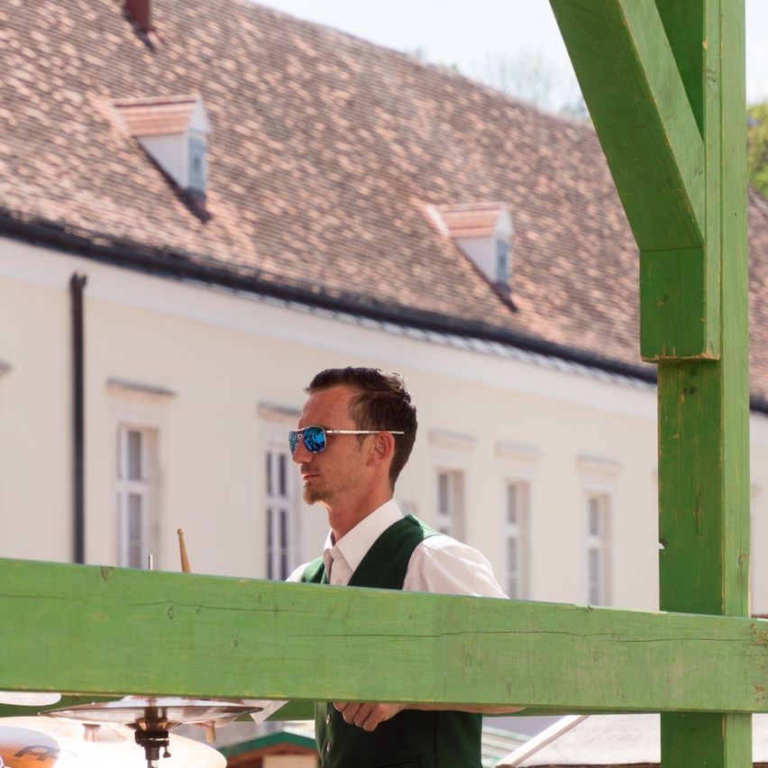 Ein Mann mit Sonnenbrille und grüner Weste steht vor einem Gebäude mit gefliestem Dach und mehreren Fenstern. Er hält Drumsticks in der Nähe eines Schlagzeugs.