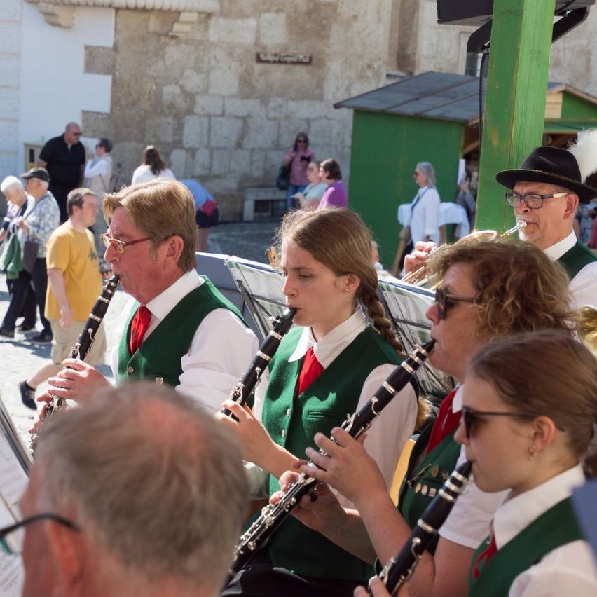 Eine Gruppe von Musikern in grünen Westen und roten Krawatten spielt Instrumente, möglicherweise Klarinetten, auf einer Kopfsteinpflasterstraße. Dahinter beobachtet ein Publikum. Im Hintergrund steht ein altes Steingebäude.