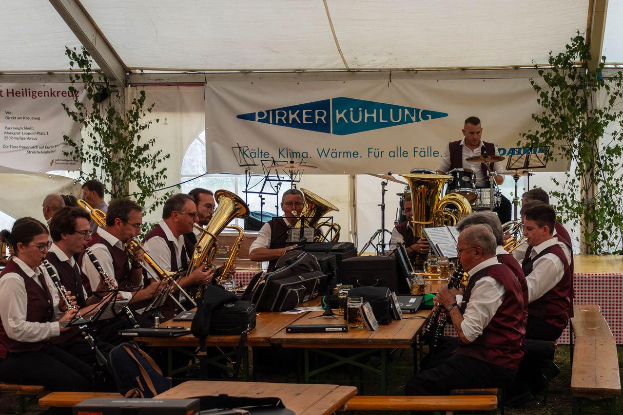 Eine Gruppe von Musikern spielt unter einem Zelt mit einem Banner, das 'Pirker Kuhlung' besagt. Sie spielen verschiedene Instrumente, darunter Tubas und Trommeln, mit ein paar Gläsern Bier auf dem Tisch.