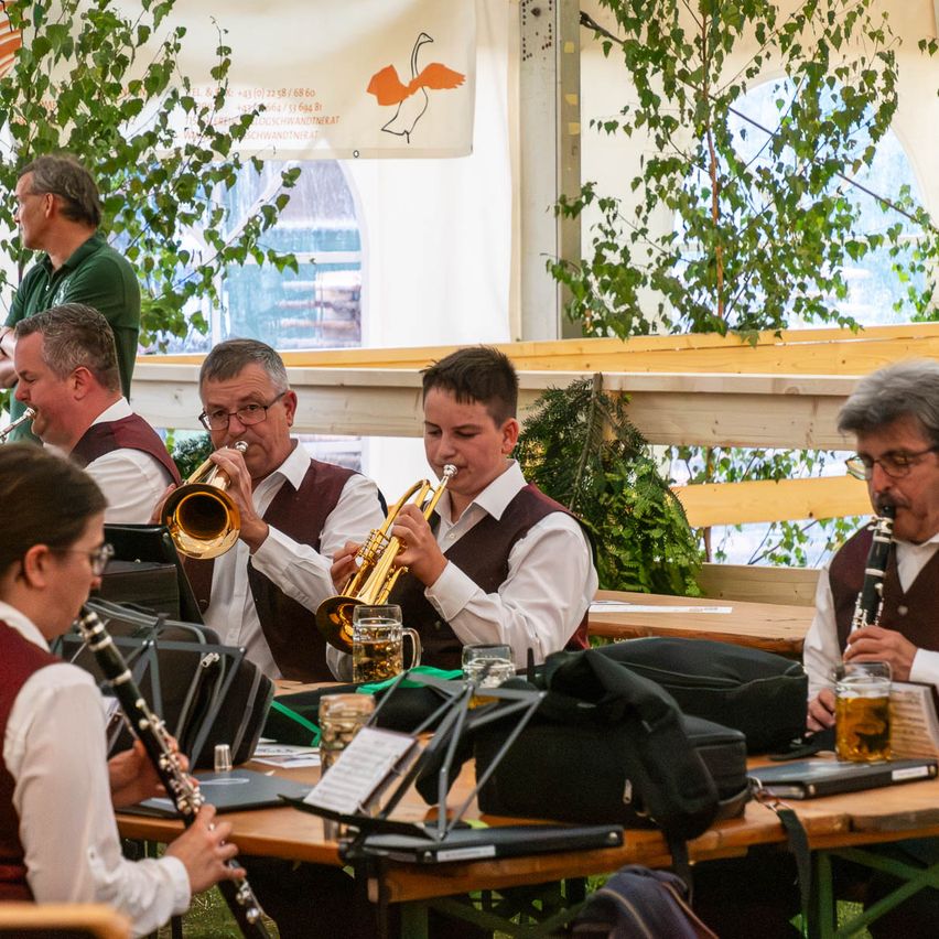 Eine Gruppe von Musikern in Westen spielt Trompeten und Klarinetten bei einer Veranstaltung im Freien, wobei ein Mann in einem grünen Shirt hinter ihnen steht.