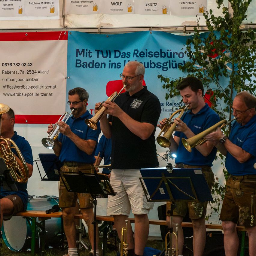 Eine Gruppe von Musikern in traditioneller Kleidung spielt Trompeten bei einer Veranstaltung im Freien. Ein Banner mit Kontaktinformationen ist im Hintergrund sichtbar.