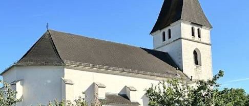 Eine weiße Kirche mit einem Schieferdach und einem Glockenturm, umgeben von Bäumen und einer Mauer an einem sonnigen Tag.