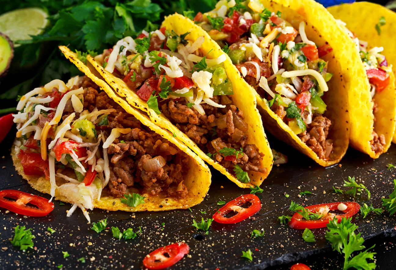 Drei Tacos mit Rindfleisch, Käse und Tomaten stehen auf einer schwarzen Oberfläche, garniert mit Petersilie und roten Chilischoten.