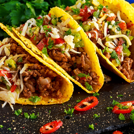 Drei Tacos mit Rindfleisch, Käse und Tomaten stehen auf einer schwarzen Oberfläche, garniert mit Petersilie und roten Chilischoten.