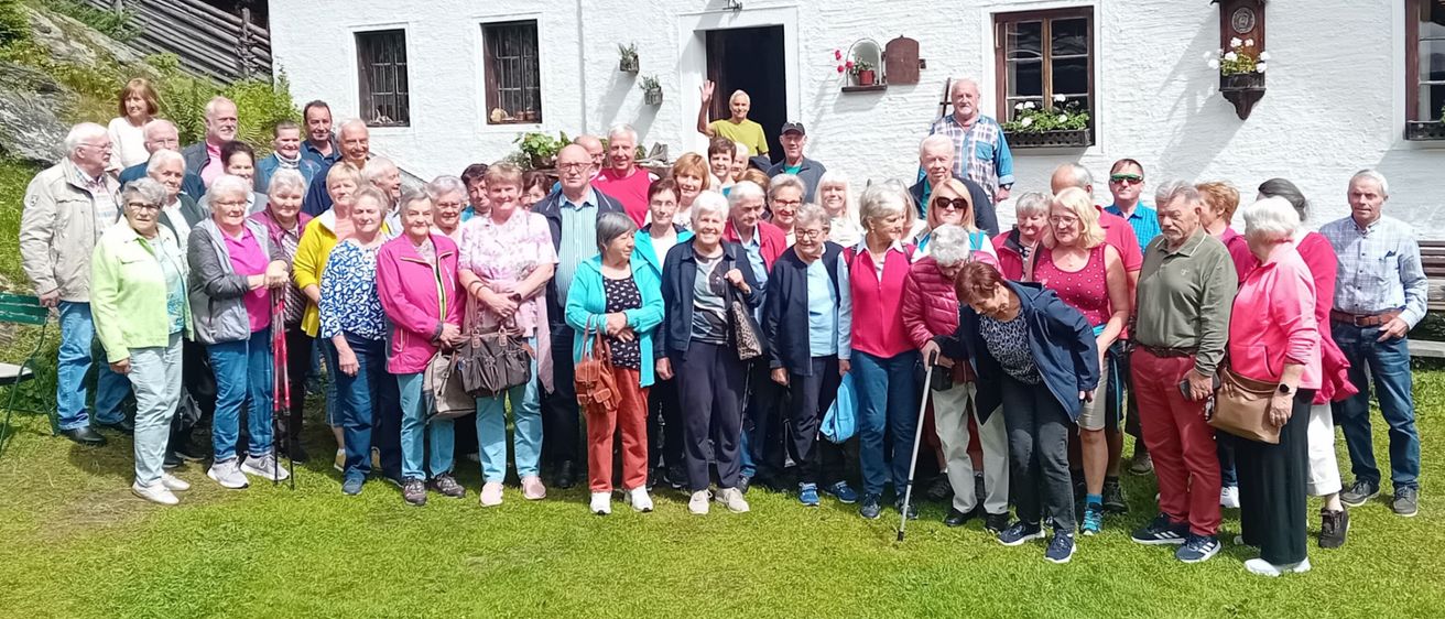 Gruppenfoto von älteren Menschen vor einem Haus mit Blumenkästen und einer Uhr an der Wand, datiert auf den 31. Juli 2025.