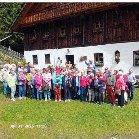 Eine Gruppe älterer Menschen steht vor einem traditionellen Holzhaus mit roten Blumen am 31. Juli 2025 um 11:20 Uhr.